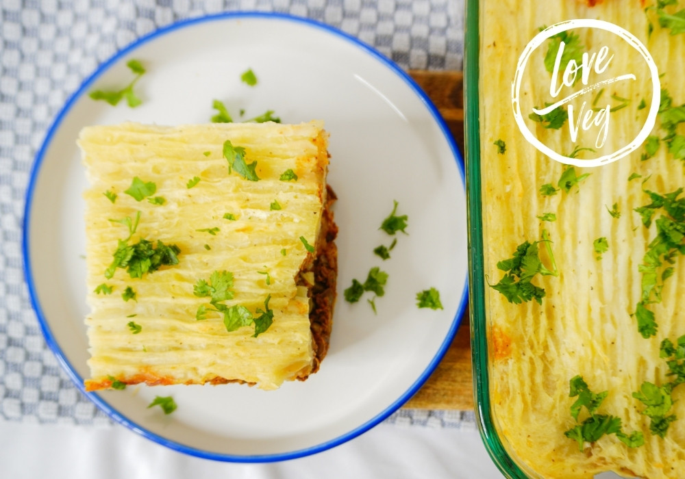 Pastel vegano en capas con puré de yuca, lentejas, champiñones y nueces, servido en un plato blanco, con la capa superior dorada y textura cremosa.