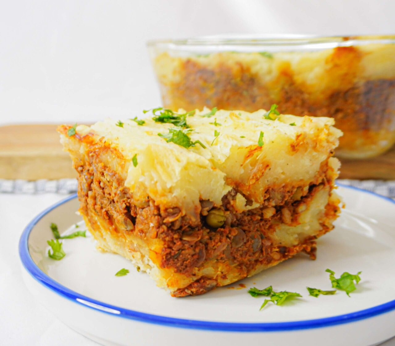 Pastel vegano en capas con puré de yuca, lentejas, champiñones y nueces, servido en un plato blanco, con la capa superior dorada y textura cremosa.