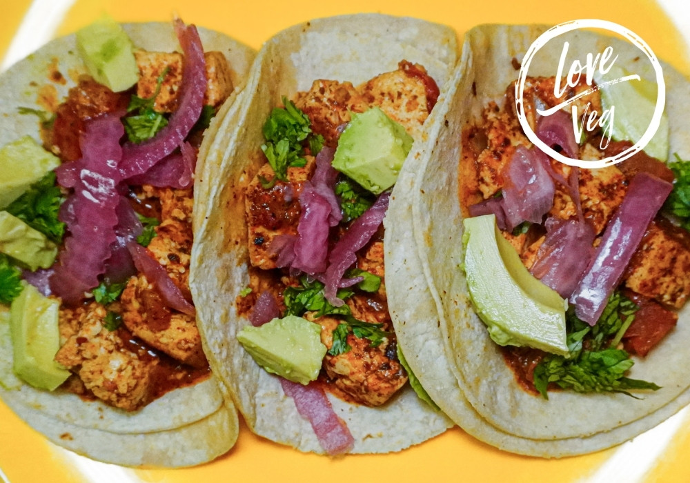 Tacos de tofu encocado sobre tortillas de maíz, servidos en un plato sobre un fondo amarillo vibrante. El tofu está bañado en una salsa cremosa de coco y achiote y decorado con cubos de aguacate fresco, cebolla morada picada y cilantro.
