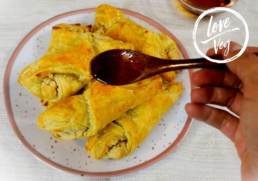 Quesitos veganos de hojaldre dorado rellenos de queso crema vegetal, servidos en un plato y bañados con miel de maple por encima.