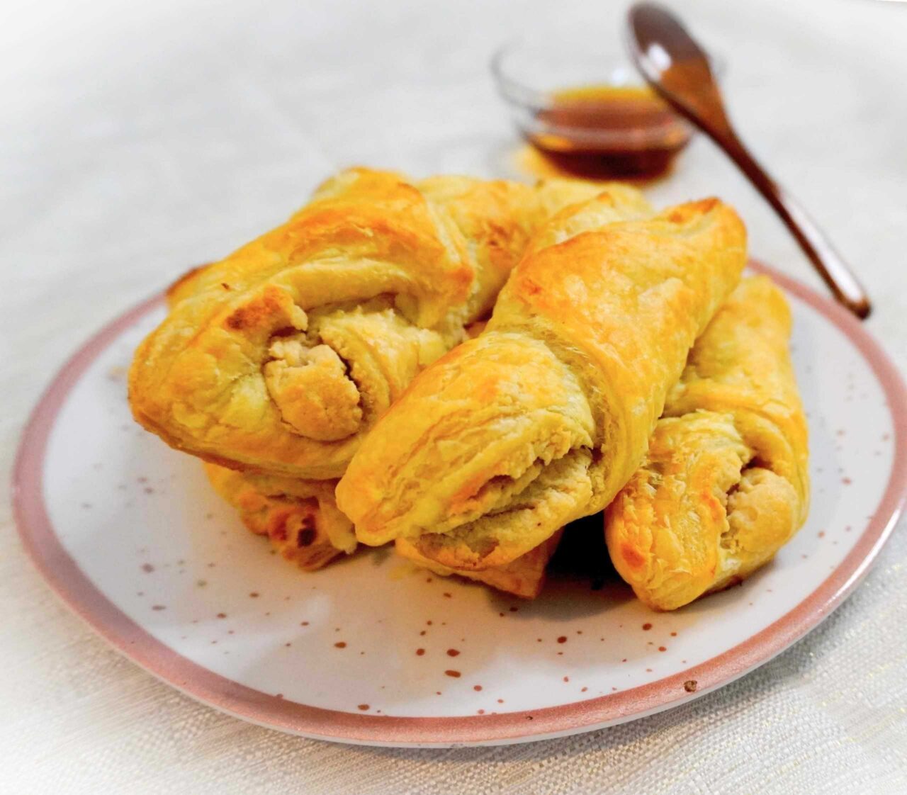 Quesitos veganos de hojaldre dorado rellenos de queso crema vegetal, servidos en un plato y bañados con miel de maple por encima.