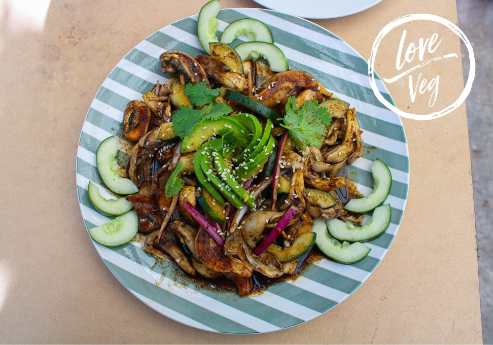 Aguachile vegetal servido en plato extendido, con hongos en tiras tipo camarón, salsa negra hecha con tortillas y chiles tatemados, rodajas de pepino y cebolla en julianas, decorado con aguacate y espolvoreado con ajonjolí, acompañado de tostadas crujientes.
