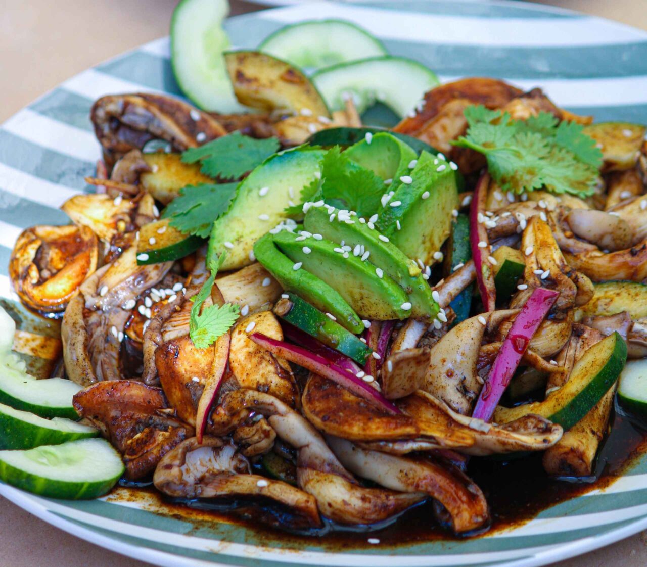 Aguachile vegetal servido en plato extendido, con hongos en tiras tipo camarón, salsa negra hecha con tortillas y chiles tatemados, rodajas de pepino y cebolla en julianas, decorado con aguacate y espolvoreado con ajonjolí, acompañado de tostadas crujientes.