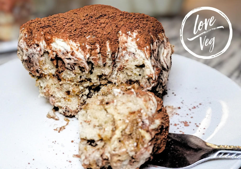 Rebanada de tiramisú vegano con capas de crema mascarpone de soya y anacardos, dedos de señora caseros y cacao en polvo sobre plato blanco.