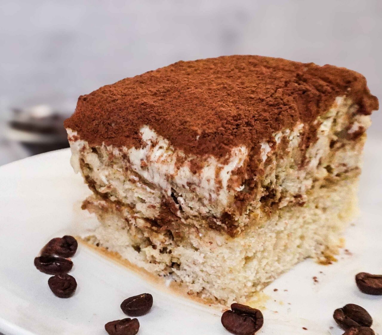 Rebanada de tiramisú vegano con capas de crema mascarpone de soya y anacardos, dedos de señora caseros y cacao en polvo sobre plato blanco.