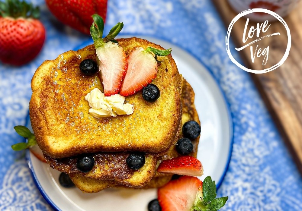 Rebanadas de tostadas francesas veganas doradas apiladas en un plato, decoradas con mantequilla derretida, arándanos frescos y rodajas de fresa sobre un mantel azul y blanco.