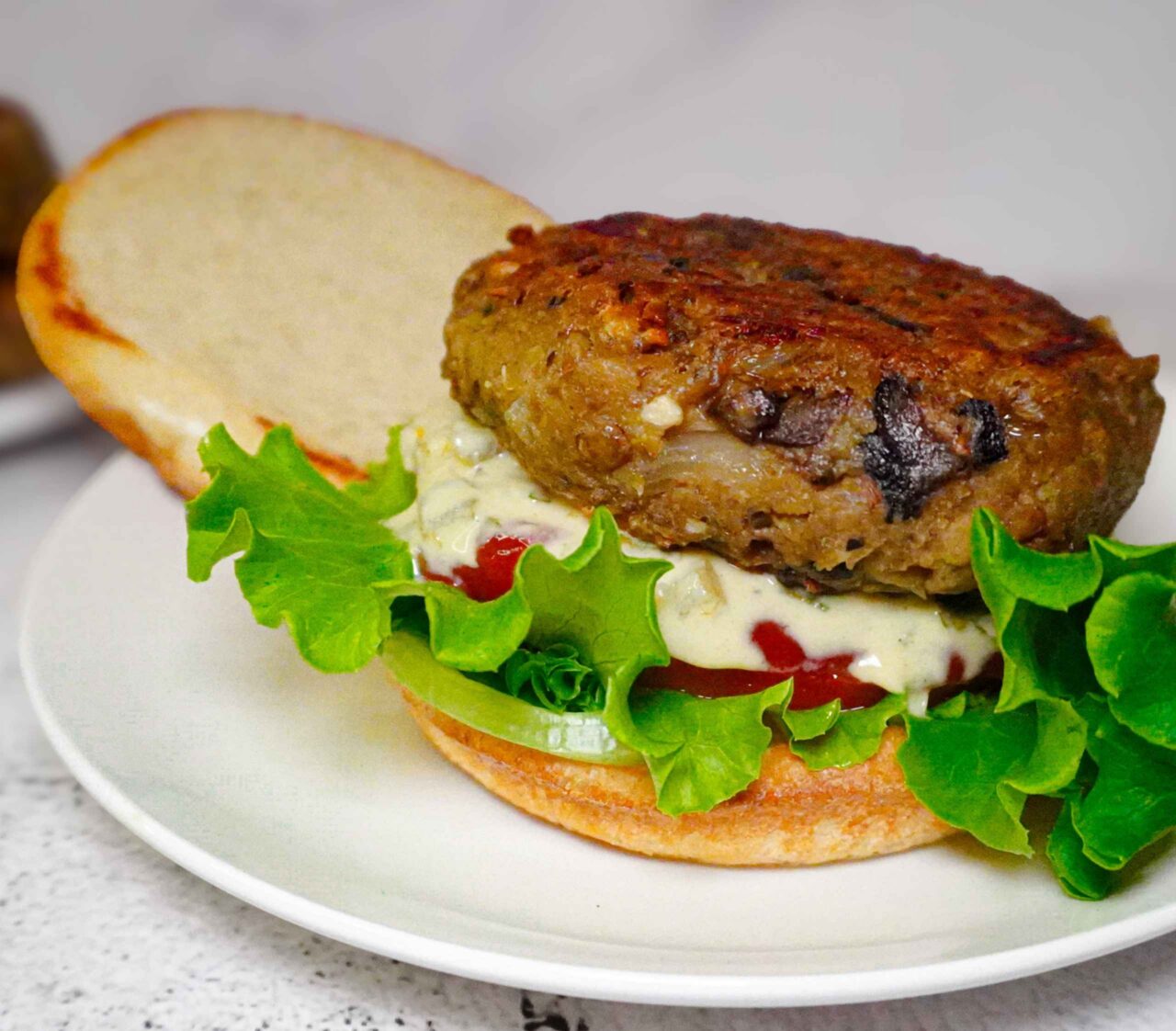 Primer plano de una hamburguesa de lentejas y champiñones vegana con costra dorada. Está servida sobre pan brioche con lechuga fresca, abundante salsa tártara cremosa con trocitos de cebolla, y una base de tomate.