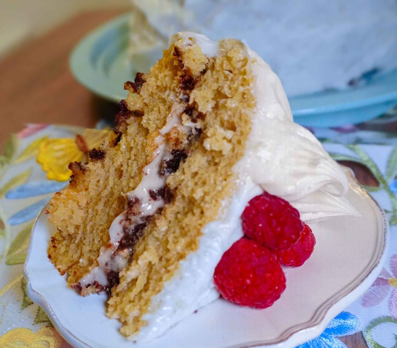 Una porción de pastel de cumpleaños vegano sobre un plato de postre decorado, con un fondo de mantel floral bordado. El pastel es esponjoso, con dos capas de bizcocho de vainilla, relleno de una crema ligera con trocitos de chocolate y cubierto con glaseado blanco. Se sirve acompañado de frambuesas frescas.