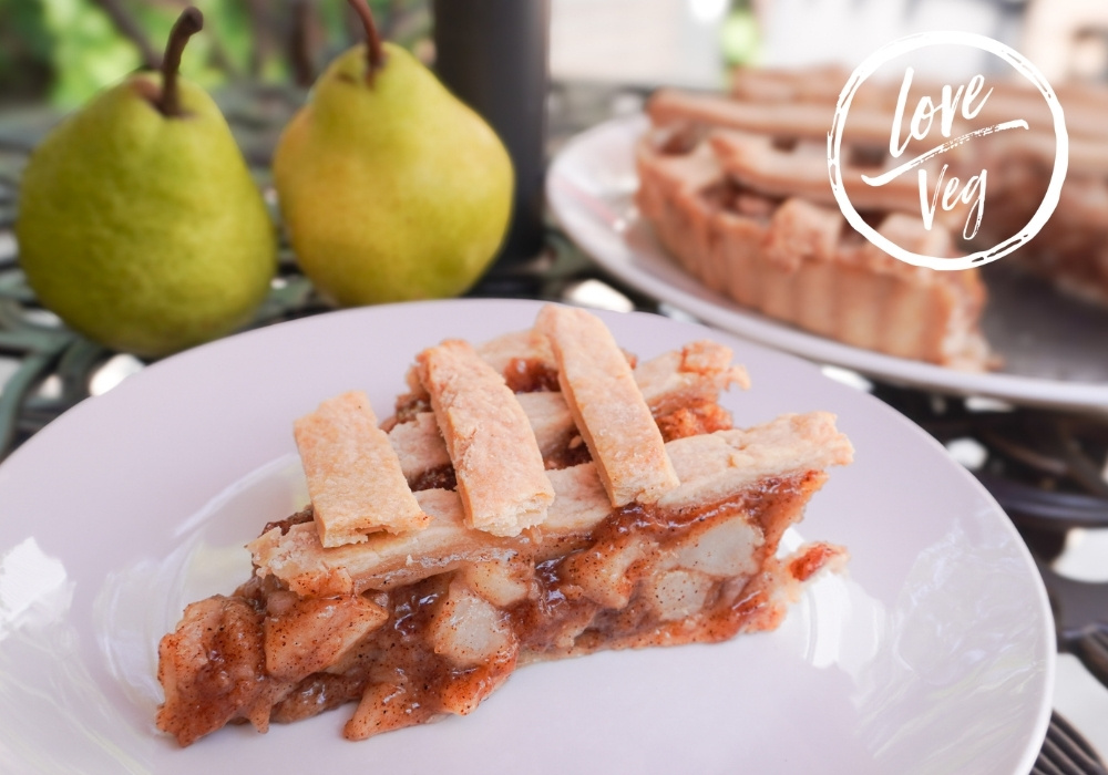 Porción de pie de pera vegano con enrejado de masa en un plato blanco, con peras frescas y la tarta completa al fondo.